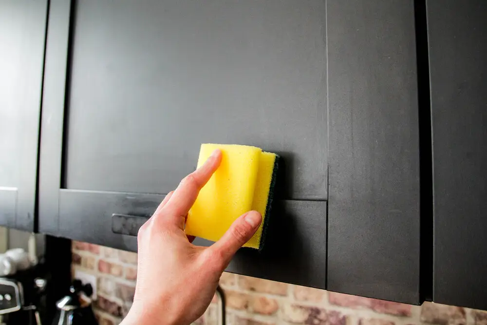 Hand wiping painted kitchen cabinets with a yellow sponge while cleaning grease and stains, demonstrating how to clean painted kitchen cabinets.